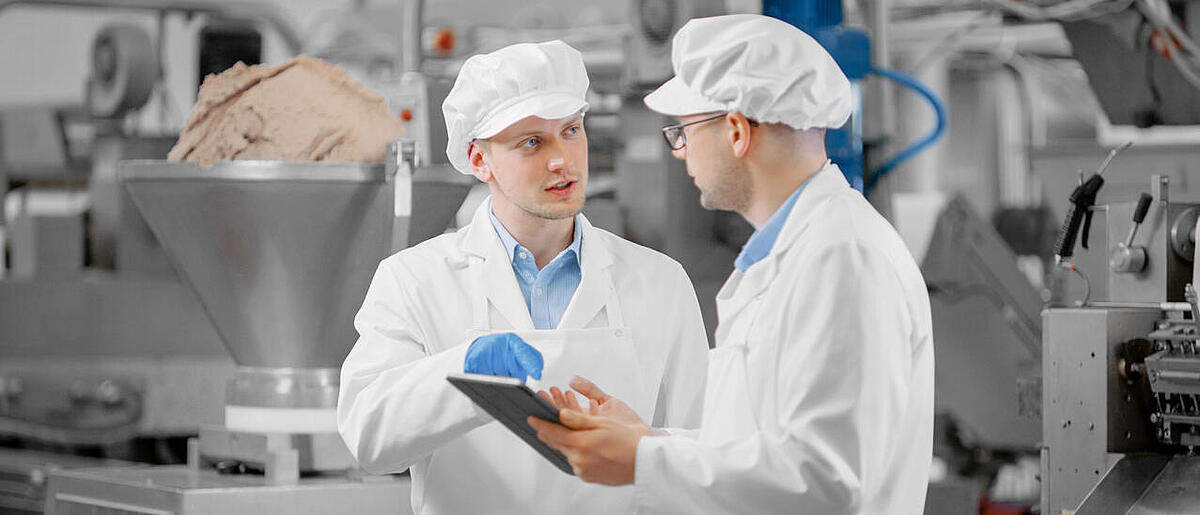 Zwei Mitarbeiter im Produktionsraum beschäftigen sich mit Arbeitsfragen. Männlicher Techniker oder Qualitätsmanager verwendet einen Tablet-Computer für die Arbeit. Sie tragen einen weißen Sanitärhut und Arbeitshandschuhe..