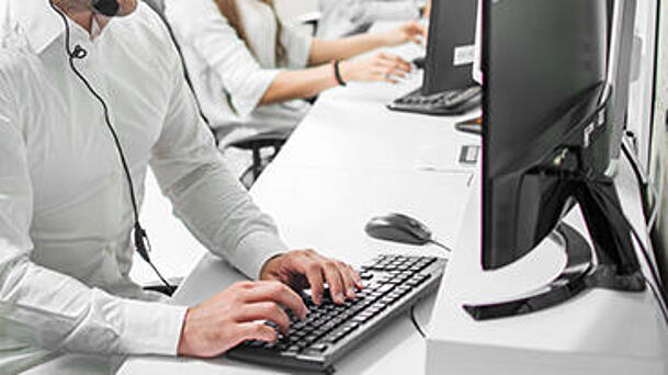 Callcenter Mitarbeiter mit Headset sitzt lächelnd am PC während er ein Gespräch mit einem Kunden führt. Im Hintergrund sitzen seine Kollegen und üben die gleiche Tätigkeit aus